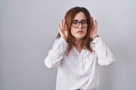 Téléchargez les photos : Femme brune debout sur fond blanc isolé essayant d'entendre les deux mains sur le geste de l'oreille, curieuse de commérages. problème auditif, sourd - en image libre de droit