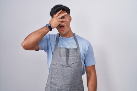 Photo for Hispanic young man wearing apron over white background peeking in shock covering face and eyes with hand, looking through fingers with embarrassed expression. - Royalty Free Image