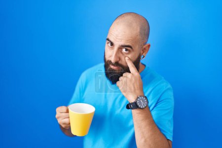 Téléchargez les photos : Jeune homme hispanique avec barbe et tatouages boire une tasse de café pointant vers l'œil vous regarder geste, expression suspecte - en image libre de droit