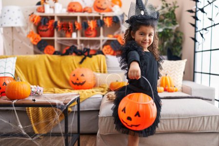 Téléchargez les photos : Adorable fille hispanique ayant fête d'Halloween tenant panier de citrouille à la maison - en image libre de droit