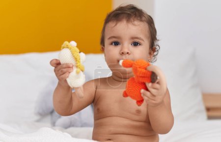 Foto de Adorable bebé hispano sentado en la cama jugando con juguetes en el dormitorio - Imagen libre de derechos