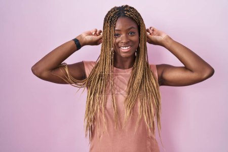 Téléchargez les photos : African american woman with braided hair standing over pink background smiling pulling ears with fingers, funny gesture. audition problem - en image libre de droit