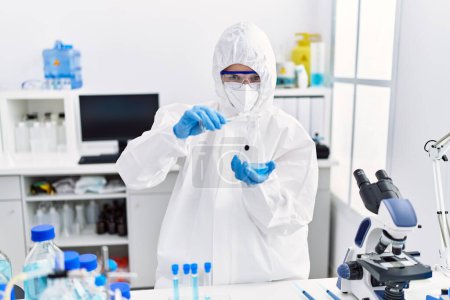 Téléchargez les photos : Jeune scientifique blonde portant un uniforme de sécurité versant du liquide sur une éprouvette au laboratoire - en image libre de droit