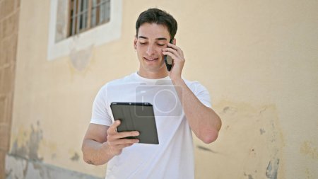 Photo for Young hispanic man talking on smartphone using touchpad at street - Royalty Free Image