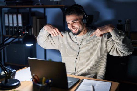 Téléchargez les photos : Jeune homme beau travaillant à l'aide d'un ordinateur portable la nuit regardant confiant avec le sourire sur le visage, se montrant du doigt fier et heureux. - en image libre de droit