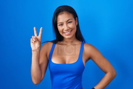 Téléchargez les photos : Femme hispanique debout sur fond bleu souriant en regardant la caméra montrant des doigts faisant signe de victoire. numéro deux. - en image libre de droit