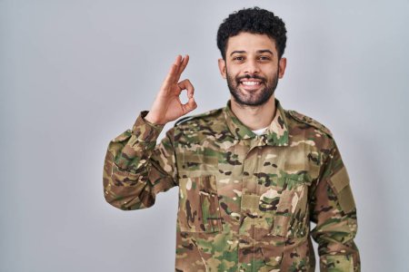 Foto de Hombre árabe vistiendo camuflaje uniforme del ejército sonriendo positiva haciendo signo de ok con la mano y los dedos. expresión exitosa. - Imagen libre de derechos