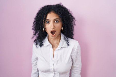 Foto de Hispanic woman with curly hair standing over pink background afraid and shocked with surprise and amazed expression, fear and excited face. - Imagen libre de derechos