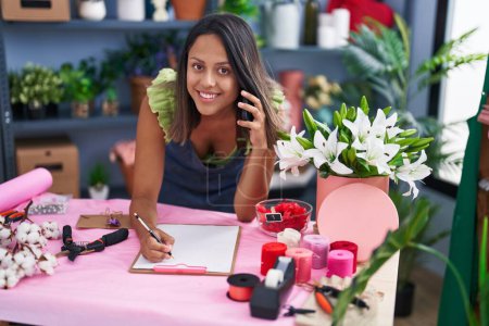 Téléchargez les photos : Jeune hispanique femme fleuriste parler sur smartphone écriture sur document au magasin de fleuriste - en image libre de droit