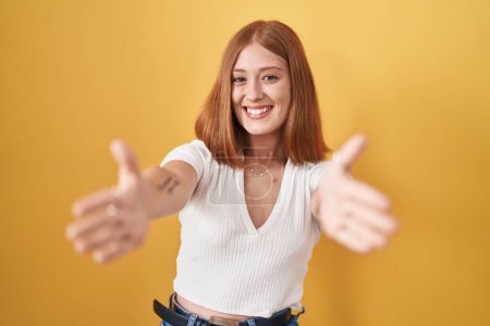 Photo for Young redhead woman standing over yellow background looking at the camera smiling with open arms for hug. cheerful expression embracing happiness. - Royalty Free Image