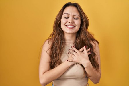 Téléchargez les photos : Jeune femme hispanique debout sur fond jaune souriant avec les mains sur la poitrine avec les yeux fermés et geste reconnaissant sur le visage. concept de santé. - en image libre de droit