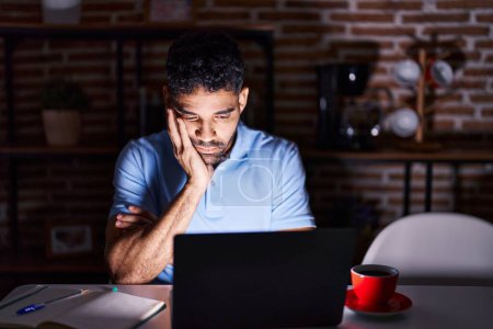 Téléchargez les photos : Homme hispanique avec barbe à l'aide d'un ordinateur portable la nuit pensant fatigué et ennuyé avec des problèmes de dépression avec les bras croisés. - en image libre de droit