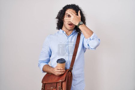 Photo for Hispanic man with curly hair drinking a cup of take away coffee peeking in shock covering face and eyes with hand, looking through fingers with embarrassed expression. - Royalty Free Image