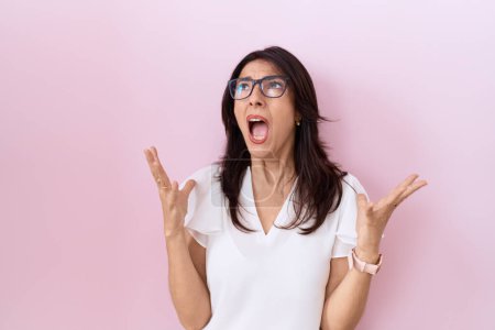 Photo for Middle age hispanic woman wearing casual white t shirt and glasses crazy and mad shouting and yelling with aggressive expression and arms raised. frustration concept. - Royalty Free Image