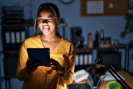 Foto de Mujer afroamericana con trenzas trabajando en la oficina por la noche con la tableta sacando la lengua feliz con expresión divertida. concepto de emoción. - Imagen libre de derechos