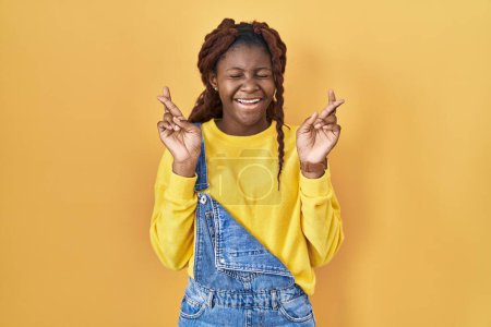 Foto de Mujer africana de pie sobre fondo amarillo gesto dedo cruzó sonriendo con esperanza y los ojos cerrados. suerte y concepto supersticioso. - Imagen libre de derechos