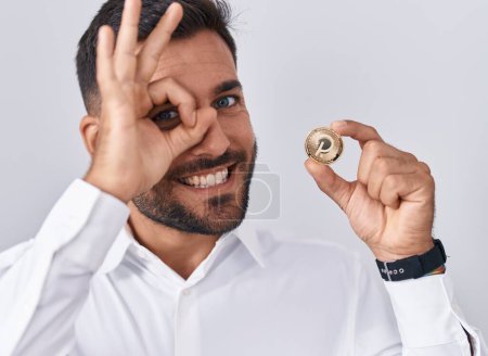 Foto de Hombre hispano guapo sosteniendo moneda criptomoneda polkadot sonriendo feliz haciendo signo ok con la mano en el ojo mirando a través de los dedos - Imagen libre de derechos