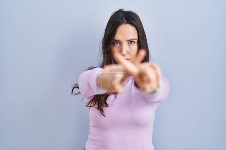 Photo for Young brunette woman standing over blue background rejection expression crossing fingers doing negative sign - Royalty Free Image