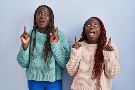 Téléchargez les photos : Deux femmes africaines debout sur fond bleu étonné et surpris regardant vers le haut et pointant avec les doigts et les bras levés. - en image libre de droit