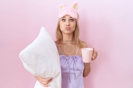 Photo for Young caucasian woman wearing pajama hugging pillow and drinking coffee puffing cheeks with funny face. mouth inflated with air, catching air. - Royalty Free Image