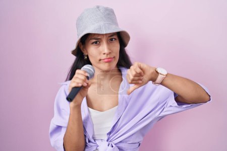 Téléchargez les photos : Jeune femme hispanique chantant chanson à l'aide d'un microphone sur fond rose avec le visage en colère, signe négatif montrant aversion avec les pouces vers le bas, concept de rejet - en image libre de droit