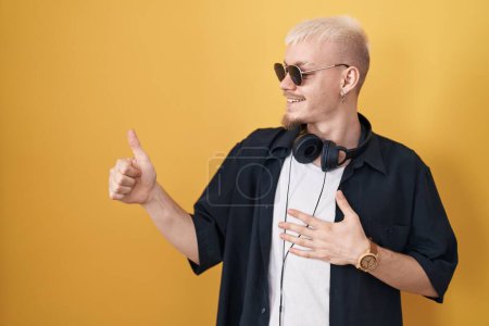 Téléchargez les photos : Jeune homme caucasien portant des lunettes de soleil debout sur fond jaune regardant fier, souriant faire les pouces vers le haut geste sur le côté - en image libre de droit