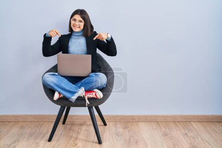 Foto de Mujer hispana joven sentada en la silla usando computadora portátil mirando confiado con sonrisa en la cara, señalándose con los dedos orgullosos y felices. - Imagen libre de derechos