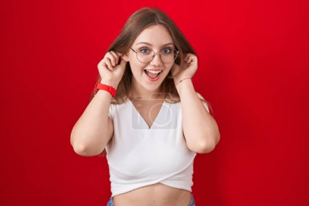Foto de Mujer caucásica joven de pie sobre fondo rojo sonriendo tirando de las orejas con los dedos, gesto divertido. problema de audición - Imagen libre de derechos