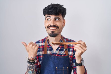 Téléchargez les photos : Jeune homme hispanique avec barbe portant tablier dégustation de nourriture tenant cuillère en bois pointant le pouce vers le côté souriant heureux avec bouche ouverte - en image libre de droit