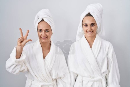Téléchargez les photos : Femme d'âge moyen et fille portant un peignoir blanc et une serviette montrant et pointant vers le haut avec les doigts numéro trois tout en souriant confiant et heureux. - en image libre de droit