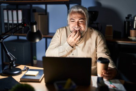 Photo for Middle age man with grey hair working at the office at night laughing and embarrassed giggle covering mouth with hands, gossip and scandal concept - Royalty Free Image