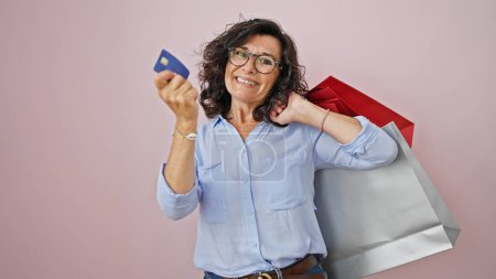 Téléchargez les photos : Moyen Age hispanique femme client tenant des sacs à provisions et carte de crédit sur fond rose isolé - en image libre de droit