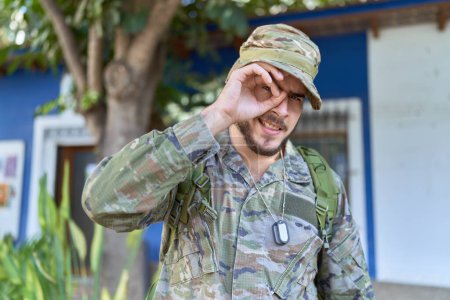 Téléchargez les photos : Jeune homme hispanique portant un uniforme de camouflage de l'armée à l'extérieur souriant heureux faisant ok signe avec la main sur les yeux regardant à travers les doigts - en image libre de droit