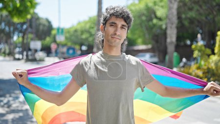 Foto de Joven hispano sosteniendo bandera de arco iris con expresión relajada en la calle - Imagen libre de derechos