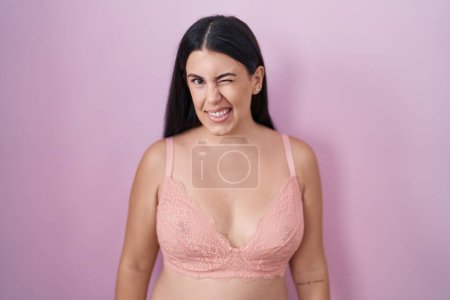 Foto de Joven mujer hispana con sujetador rosa guiñando el ojo mirando a la cámara con expresión sexy, cara alegre y feliz. - Imagen libre de derechos