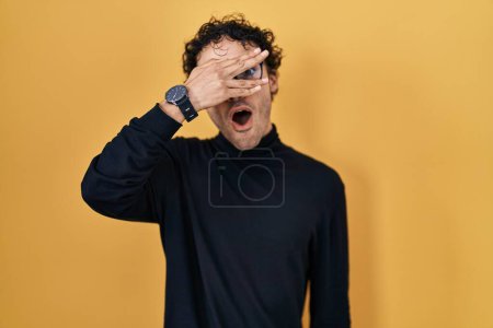 Téléchargez les photos : Homme hispanique debout sur fond jaune regardant dans le choc couvrant le visage et les yeux avec la main, regardant à travers les doigts avec une expression embarrassée. - en image libre de droit