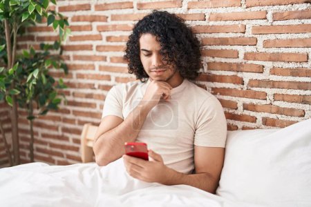 Téléchargez les photos : Homme hispanique avec les cheveux bouclés en utilisant smartphone assis sur le lit visage sérieux penser à la question avec la main sur le menton, réfléchi sur l'idée déroutante - en image libre de droit