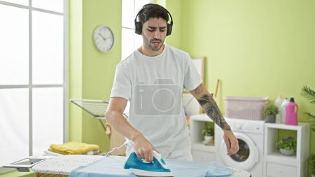 Photo for Young hispanic man listening to music ironing clothes at laundry room - Royalty Free Image