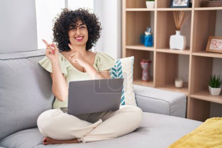 Photo for Young brunette woman with curly hair using laptop sitting on the sofa at home smiling and looking at the camera pointing with two hands and fingers to the side. - Royalty Free Image