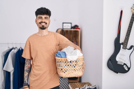 Foto de Joven hombre hispano con barba sosteniendo la cesta de la ropa en el dormitorio mirando positiva y feliz de pie y sonriendo con una sonrisa confiada mostrando los dientes - Imagen libre de derechos