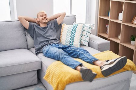 Téléchargez les photos : Jeune homme caucasien détendu avec les mains sur la tête couché sur le canapé à la maison - en image libre de droit