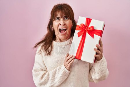 Photo for Middle age hispanic woman holding present angry and mad screaming frustrated and furious, shouting with anger looking up. - Royalty Free Image