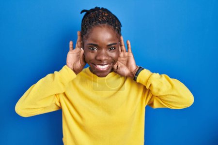 Photo for Beautiful black woman standing over blue background trying to hear both hands on ear gesture, curious for gossip. hearing problem, deaf - Royalty Free Image