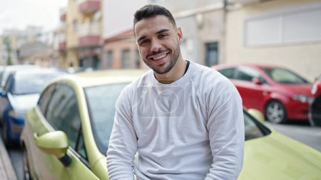 Téléchargez les photos : Jeune homme hispanique souriant confiant assis sur la voiture à la rue - en image libre de droit