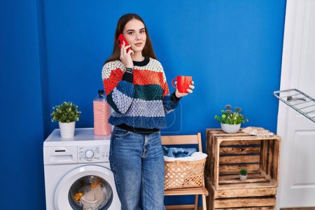 Foto de Mujer joven hablando en smartphone bebiendo café en la lavandería - Imagen libre de derechos