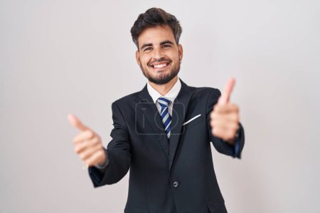 Téléchargez les photos : Jeune homme hispanique avec des tatouages portant un costume d'affaires et cravate approuvant faire un geste positif avec la main, pouces levés souriant et heureux pour le succès. geste gagnant. - en image libre de droit