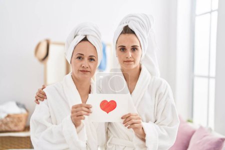 Photo for Middle age woman and daughter wearing bath robe holding heart card relaxed with serious expression on face. simple and natural looking at the camera. - Royalty Free Image