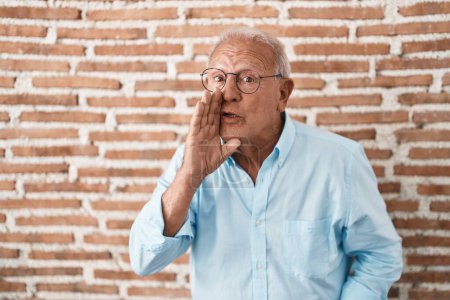 Foto de Hombre mayor con el pelo gris de pie sobre ladrillos pared mano en boca diciendo rumor secreto, susurrando conversación maliciosa - Imagen libre de derechos