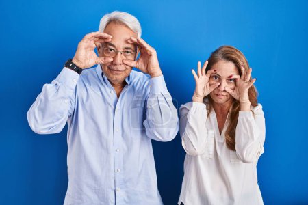 Téléchargez les photos : Couple hispanique moyen âge debout sur fond bleu essayant d'ouvrir les yeux avec les doigts, somnolent et fatigué pour la fatigue matinale - en image libre de droit