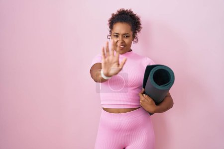 Téléchargez les photos : Jeune femme hispanique avec les cheveux bouclés tenant tapis de yoga sur fond rose faisant geste d'arrêt avec les paumes des mains, expression de colère et de frustration - en image libre de droit